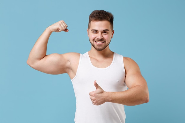 Sonriente joven barbudo de 20 años en camiseta blanca posando aislado en un retrato de estudio de fondo azul pastel. Deporte fitness concepto de estilo de vida saludable. Simulacros de espacio de copia. Mostrando músculos bíceps, pulgar hacia arriba.