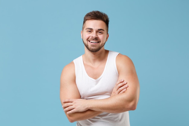 Sonriente joven barbudo de 20 años en camiseta blanca posando aislado en un retrato de estudio de fondo de pared azul pastel. Deporte fitness concepto de estilo de vida saludable. Simulacros de espacio de copia. Tomados de la mano cruzados.