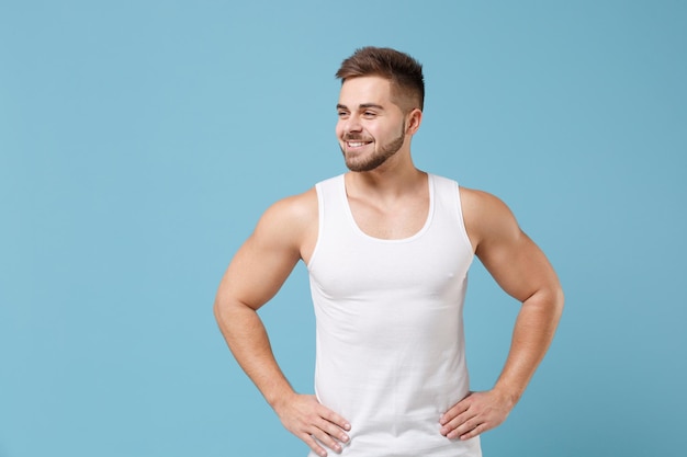 Sonriente joven barbudo de 20 años en camiseta blanca posando aislado sobre fondo azul pastel. Deporte fitness concepto de estilo de vida saludable. Simulacros de espacio de copia. De pie con los brazos en jarras sobre la cintura mirando a un lado.