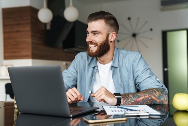 Sonriente joven barbudo que trabaja en la computadora portátil mientras está sentado en la mesa de la cocina