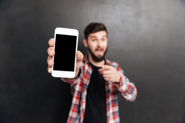 Foto sonriente joven barbudo sosteniendo y mostrando el teléfono celular de pantalla en blanco en gris