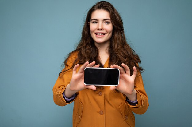 Sonriente joven y bella mujer morena con chaqueta naranja aislada sobre fondo azul sosteniendo