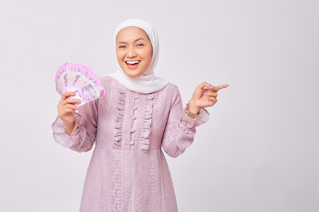 Sonriente joven y bella mujer musulmana asiática con hiyab y vestido morado sosteniendo billetes de dinero en efectivo en rupias y señalando con el dedo el espacio vacío aislado en el fondo blanco del estudio