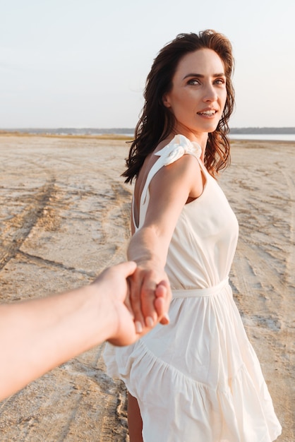 Sonriente joven y bella mujer con vestido de verano sosteniendo a sus novios con la mano extendida mientras camina en la playa