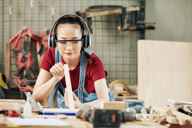 Sonriente joven carpintero en orejeras y gafas comprobando la uniformidad en la plancha de madera que cortó