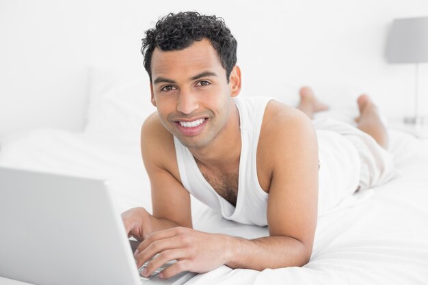 Sonriente joven casual con laptop en la cama