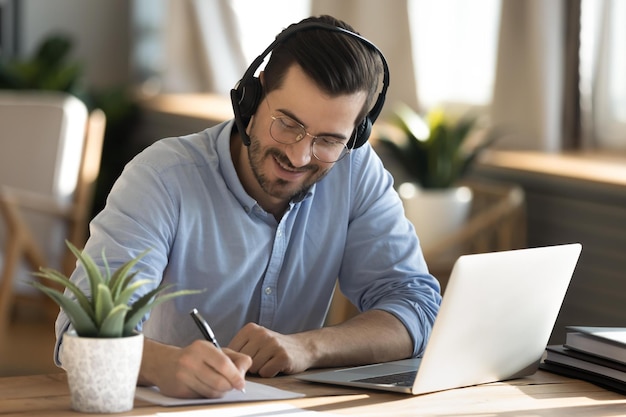 Foto sonriente joven caucásico con gafas de auriculares sentado en el escritorio de trabajo en la computadora portátil haciendo notas feliz hombre milenario en auriculares mira seminario web o curso de capacitación o estudio de computadora en línea desde casa