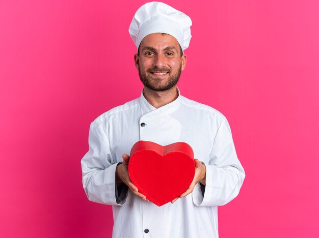 Sonriente joven cocinero masculino caucásico en uniforme de chef y gorra con forma de corazón