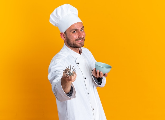 Sonriente joven cocinero de sexo masculino caucásico en uniforme de chef y gorra de pie en la vista de perfil sosteniendo el cuenco mirando y apuntando a la cámara con un batidor aislado en la pared naranja con espacio de copia