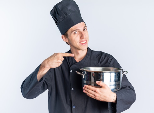 Sonriente joven cocinero de sexo masculino rubio en uniforme de chef y gorra sosteniendo y apuntando a la olla