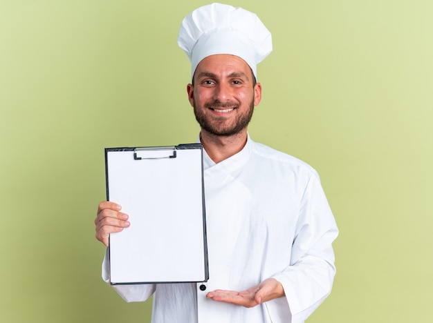 Sonriente joven cocinero varón caucásico en uniforme de chef y gorra mostrando portapapeles apuntando