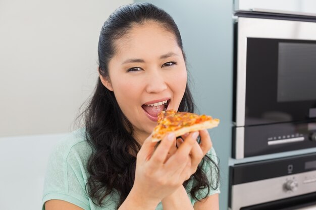 Sonriente joven comiendo una rebanada de pizza en la cocina