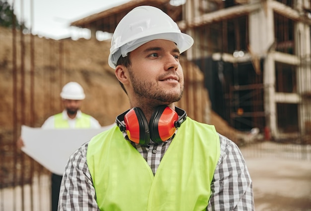 Foto sonriente joven contratista de pie en el sitio de construcción y mirando a otro lado