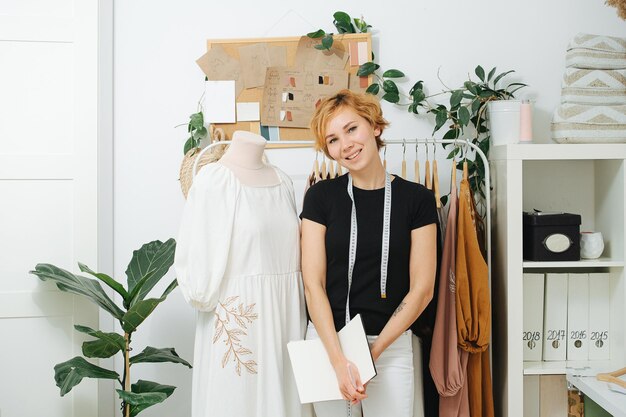 Sonriente joven costurera frente a un estante junto a un maniquí con un vestido blanco Tiene el pelo corto teñido de jengibre Medidor colgando de su cuello