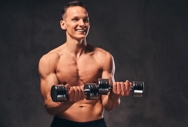 Sonriente joven culturista sin camisa con cuerpo musculoso haciendo ejercicio con pesas sobre un fondo oscuro.