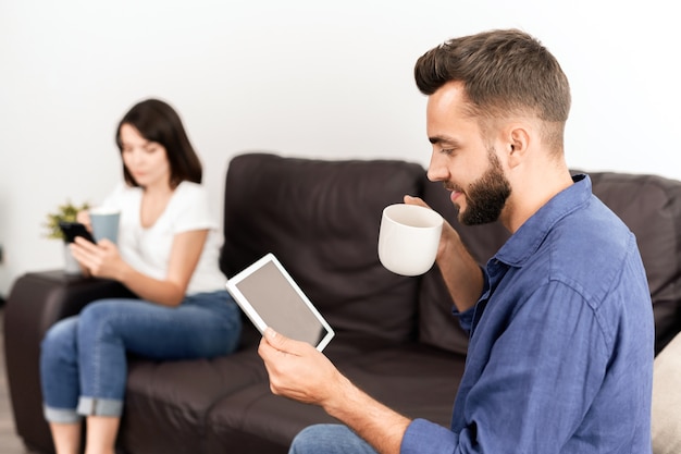 Sonriente joven curioso en camisa casual sentado en el sofá con novia adicta al teléfono inteligente y leyendo noticias en línea en tableta mientras toma café