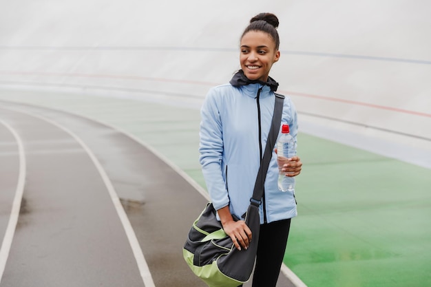 Sonriente joven deportista africana en forma con bolsa de deporte de pie en el estadio, sosteniendo una botella de agua