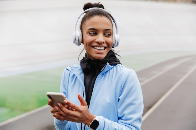 Sonriente joven deportista africana sana escuchando música con auriculares inalámbricos escurriendo en el estadio, sosteniendo el teléfono móvil