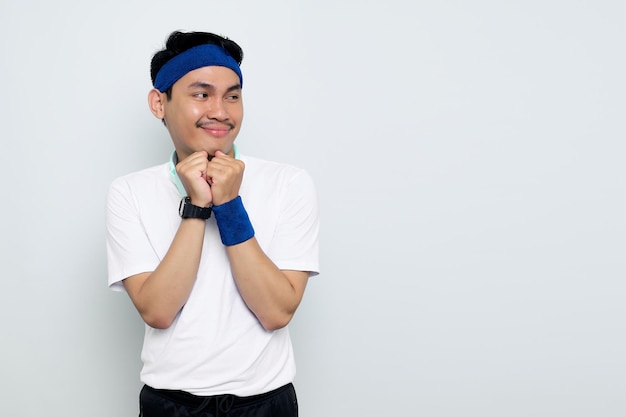 Sonriente joven deportista asiático en diadema azul y camiseta blanca con auriculares mantiene las manos debajo de la barbilla mira a un lado recuerda momento agradable aislado sobre fondo blanco Concepto de deporte de entrenamiento