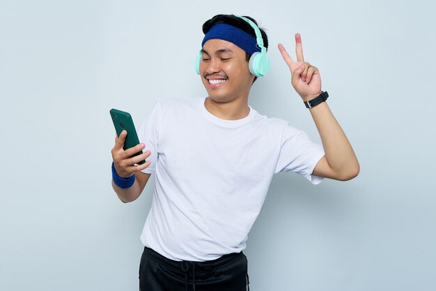 Sonriente joven deportista asiático con diadema azul y camiseta blanca de ropa deportiva mientras escucha música favorita con auriculares haciendo selfie en gesto de paz de teléfono móvil aislado en fondo blanco