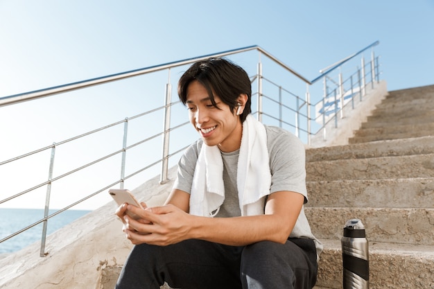 Sonriente joven deportista asiático sentado en las escaleras a la orilla del mar, enviando mensajes de texto en el teléfono móvil mientras descansa