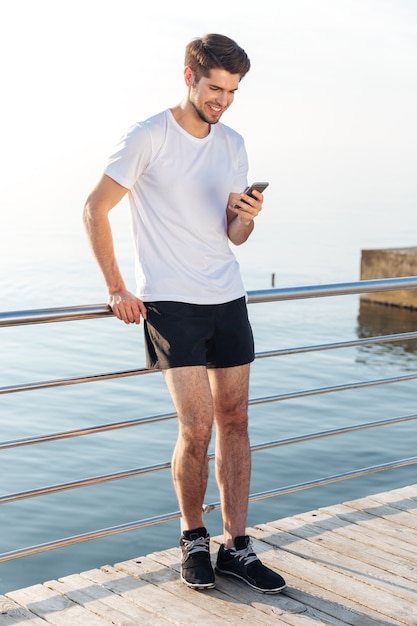 Sonriente joven deportista de pie en la terraza de madera cerca del mar y con smartphone