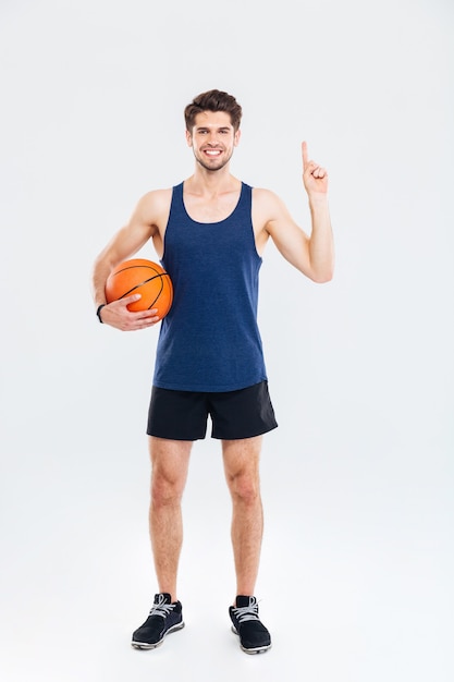 Sonriente joven deportista sosteniendo baloncesto y apuntando con el dedo hacia arriba en copyspace aislado sobre un fondo gris