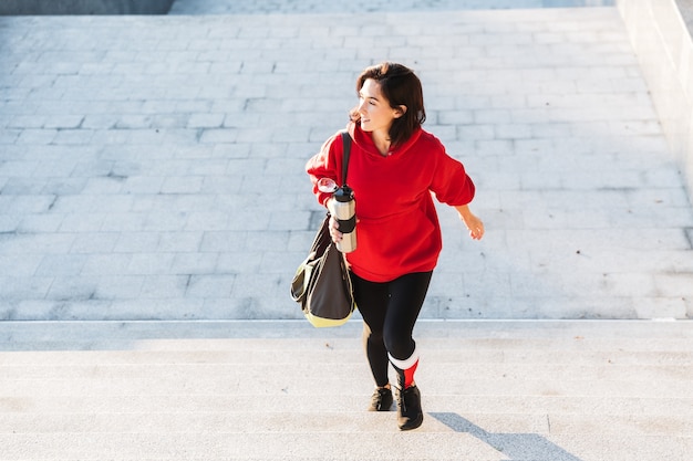 Sonriente joven deportista vistiendo una sudadera con capucha caminando al aire libre, llevando bolsa de deportes, sosteniendo la taza de café