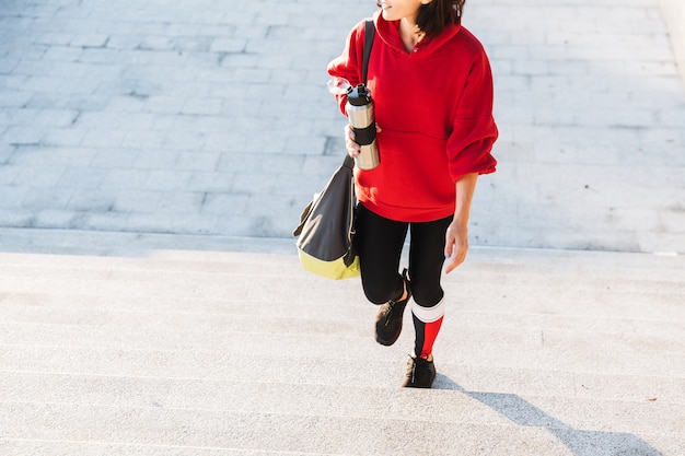 Sonriente joven deportista vistiendo una sudadera con capucha caminando al aire libre, llevando bolsa de deportes, sosteniendo la taza de café