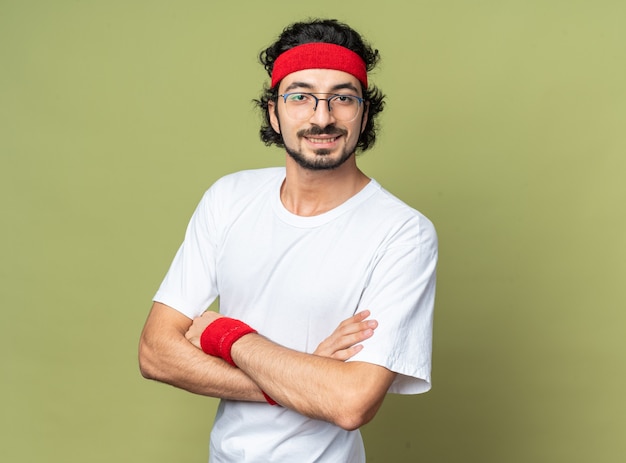 Sonriente joven deportivo vistiendo diadema con muñequera cruzando las manos