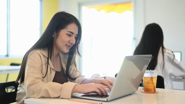 Sonriente joven diseñadora que trabaja en la computadora portátil mientras está sentada con su colega en la oficina creativa.