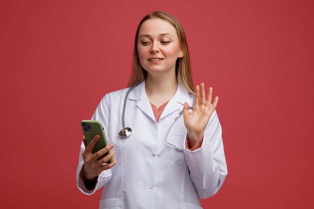 Sonriente joven doctora rubia vistiendo bata médica y estetoscopio sosteniendo y mirando el teléfono móvil agitando alrededor del cuello