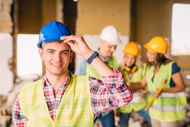 Foto sonriente joven electricista sosteniendo casco y mirando a cámara. sus colegas arquitectos revisan el plan en segundo plano en un sitio de construcción.