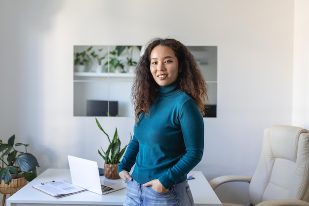 Sonriente joven empleada asiática parada en el escritorio en la oficina mira a distancia pensando o visualizando el éxito profesional Feliz plan o sueño de empresaria en el lugar de trabajo Concepto de visión empresarial