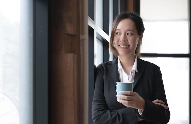 Sonriente joven empresaria asiática sosteniendo una taza de café en la oficina