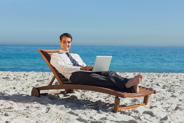 Sonriente joven empresario acostado en una silla de cubierta con su computadora portátil