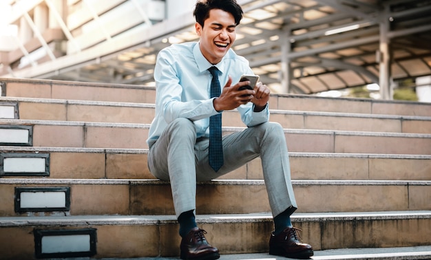 Sonriente joven empresario asiático mediante teléfono móvil en la ciudad. Sentado en la escalera