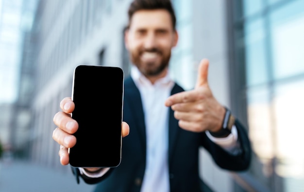 Sonriente joven empresario barbudo europeo en traje se regocija por el éxito y señala con el dedo al teléfono inteligente