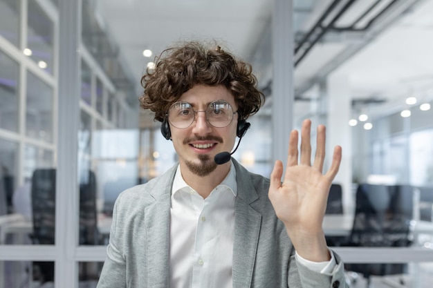 Sonriente joven empresario con el cabello rizado y auriculares agitando en la oficina que representa un amistoso