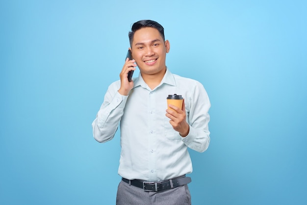 Sonriente joven empresario guapo hablando por teléfono inteligente y mantenga una taza de café sobre fondo azul.