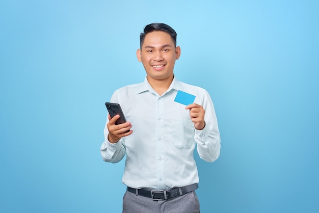 Sonriente joven empresario guapo sosteniendo smartphone y tarjeta de crédito sobre fondo azul.