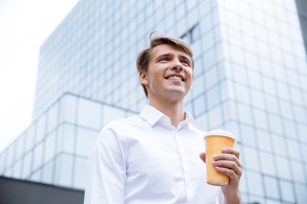 Sonriente joven empresario de pie cerca del centro de negocios y tomando café