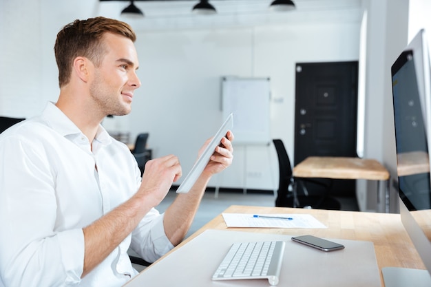 Sonriente joven empresario con tableta y computadora en el lugar de trabajo