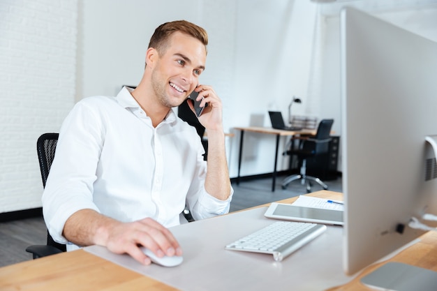 Sonriente joven empresario trabajando con la computadora y hablando por teléfono celular en el lugar de trabajo