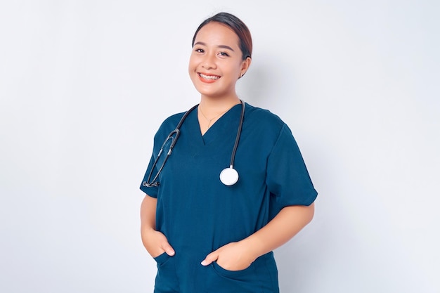 Sonriente joven enfermera asiática vistiendo uniforme azul con estetoscopio cogidos de la mano en bolsillos aislados sobre fondo blanco Concepto de medicina sanitaria