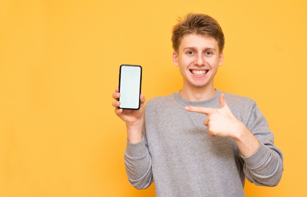 Sonriente joven está parado en amarillo y muestra un dedo en un teléfono inteligente