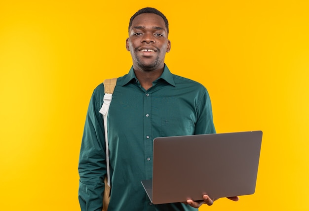 Sonriente joven estudiante afroamericano con mochila sosteniendo portátil aislado en pared naranja con espacio de copia