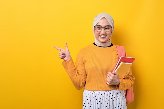 Sonriente joven estudiante asiática con mochila sosteniendo un cuaderno señalando con el dedo el espacio de copia aislado sobre fondo amarillo