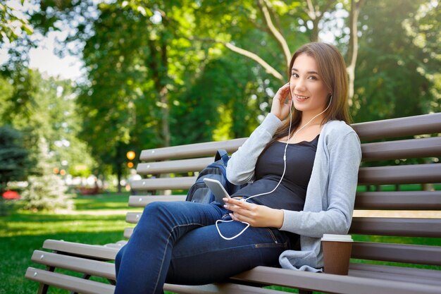 Sonriente joven estudiante en auriculares. Relajarse y escuchar música, mientras toma café en el parque. Concepto de estilo de vida y tecnología.