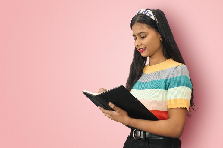 Sonriente joven estudiante india asiática posando aislada de lado haciendo notas en el bloc de notas con bolígrafo mirando hacia el bloc de notas con espacio de copia de maqueta